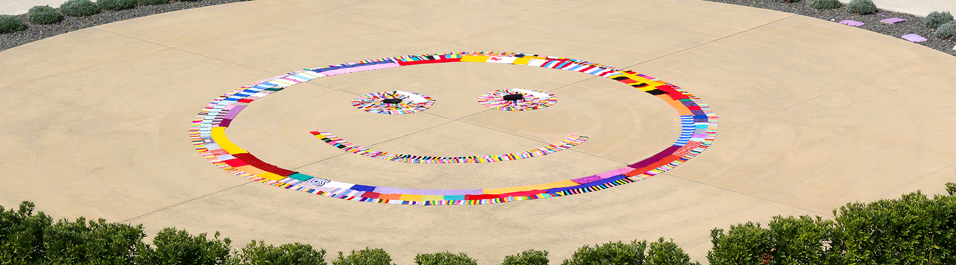 The Labyrinth Garden, decorated with colourful Warm Trees scarves forming a large smiley face. The Labyrinth Garden is bordered by neatly trimmed hedges.