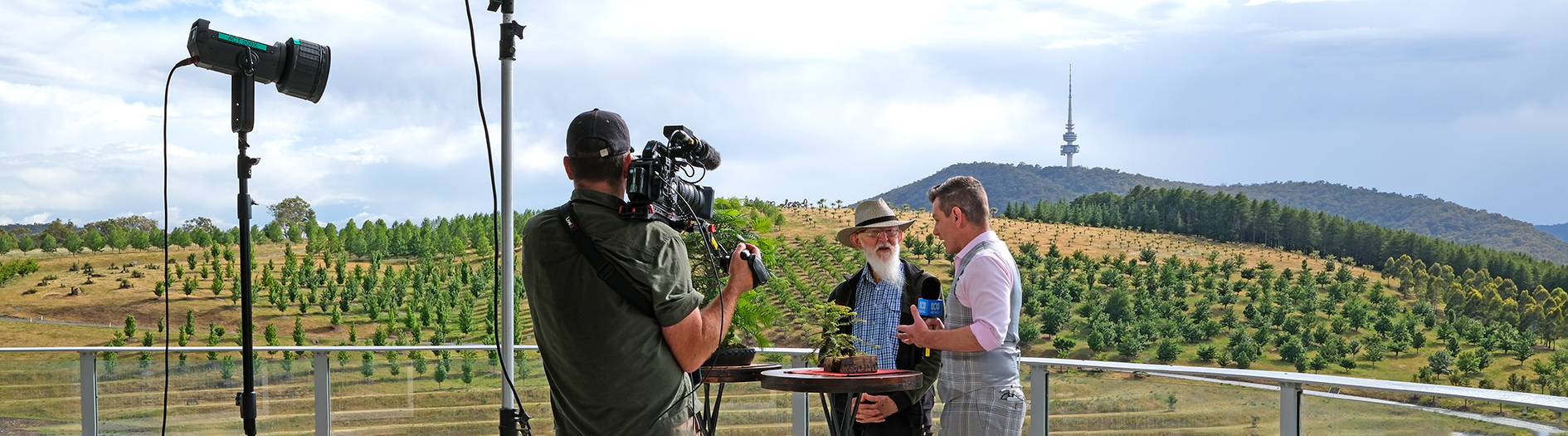 Volunteer Roger Hnatuik stands in front of a small camera crew with sound and lighting equipment, with the arboretum forests a beautiful green in the morning sun