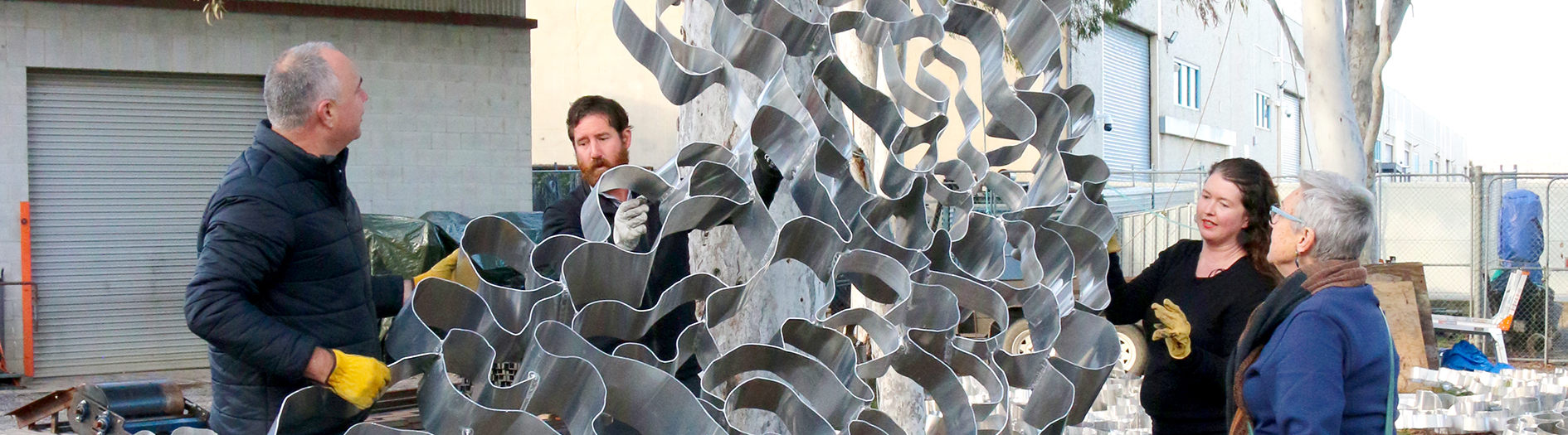 The twisted metal detail of Hannahah Quinliven's piece 'Drawing Breath' leant against a tree with Hannah speaking to the piece to members attending the visit.