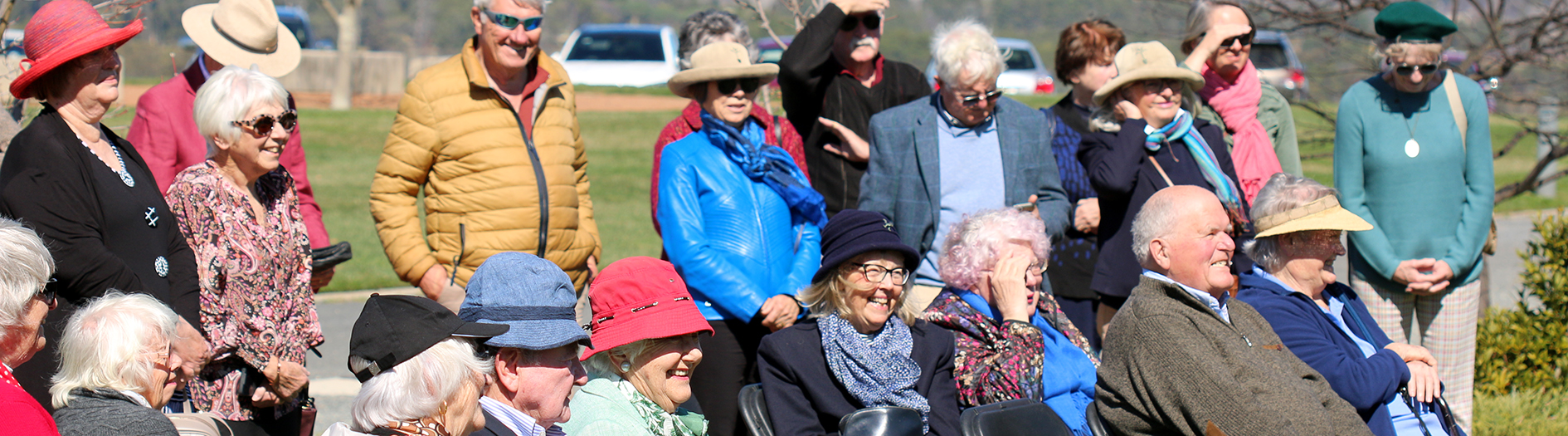A group of adults gathered in a garden, both seated and standing, all smiling on a sunny day