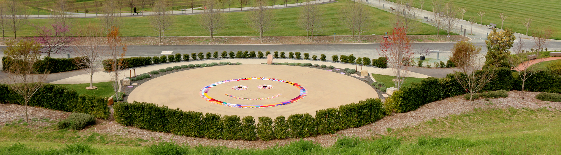 The Gallery of Gardens featuring the Labyrinth Garden the foreground, decorated with colourful Warm Trees scarves forming a large smiley face. The Labyrinth Garden is bordered by neatly trimmed hedges and greenery. In the background, the Events Terrace pathways lead to the Margaret Whitlam Pavilion, with hills and distant buildings of Canberra visible on the horizon.