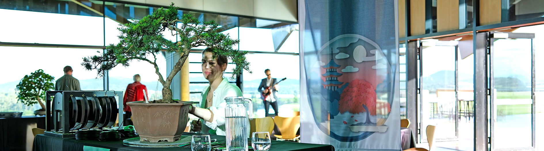 Bonsai artists demonstrates her art on stage, with a musician visible in the background
