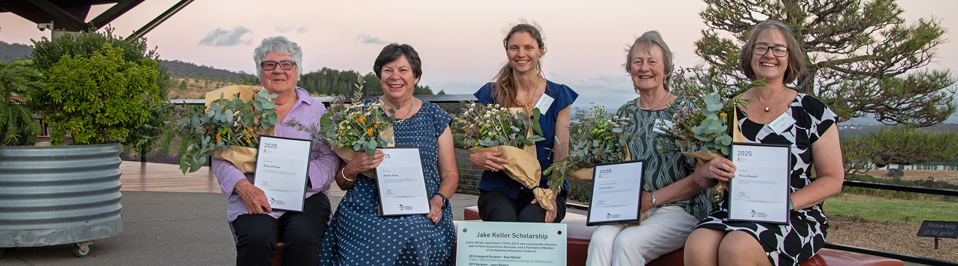 Award recipients gather outside in a garden, each holding their awards and a bouquet of flowers, with a purple dusky sunset sky behind them