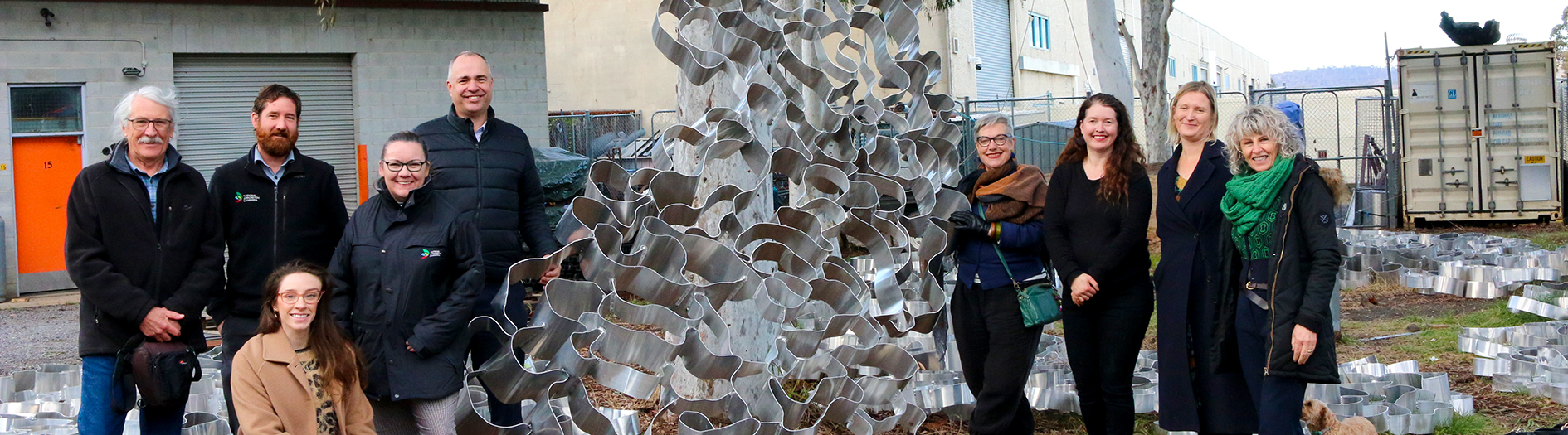 The twisted metal detail of Hannahah Quinliven's piece 'Drawing Breath' leant against a tree with a group photo of members attending the studio visit.