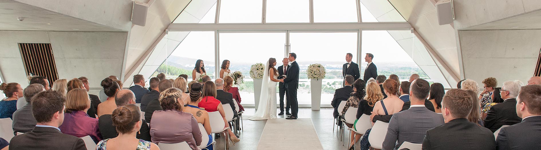 Bride and groom standing with a celebrant as their wedding guests look on, in the Margaret Whitlam Pavilion
