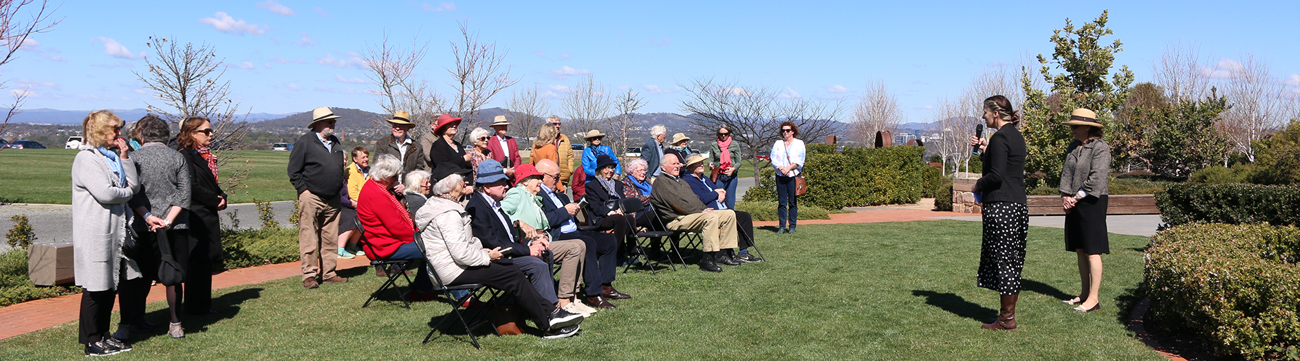 A crowd of approximately 30 adults gather on chairs listening to female presenter on a sunny day