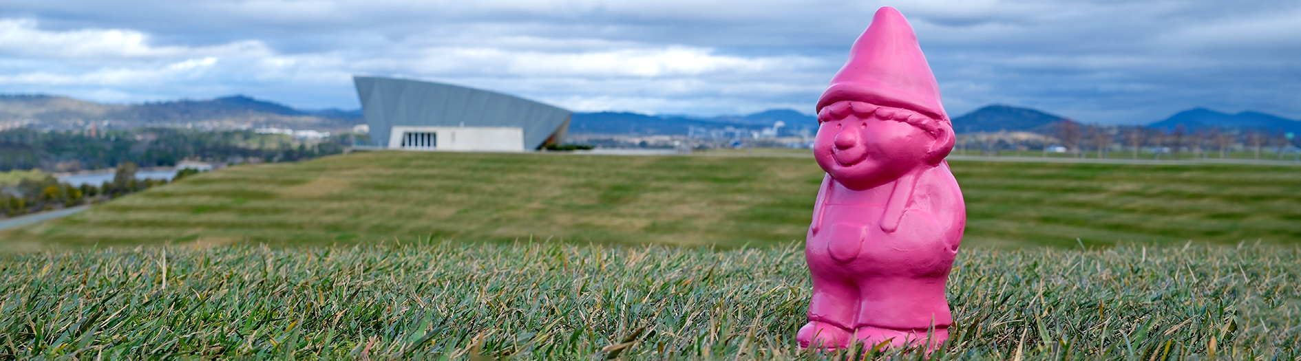 Photo of the gnome standing in the grass at the top of the amphitheatre, with the Margaret Whitelam Pavilion in the background and pink sunset clouds.