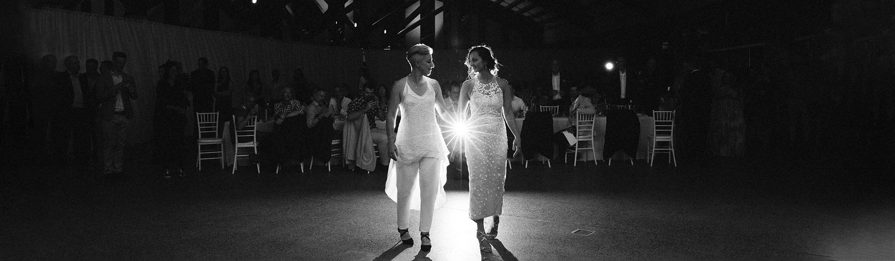 Black and white photo of two brides about to start dancing at their wedding in the Visitors Centre with low lighting and spotlights focused on the brides.