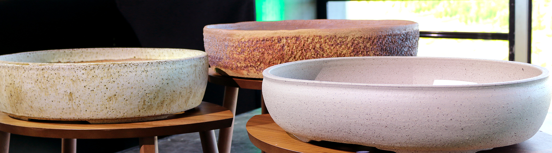 Three large, shallow, round ceramic pots, each a different colour and texture are displayed on wooden stools