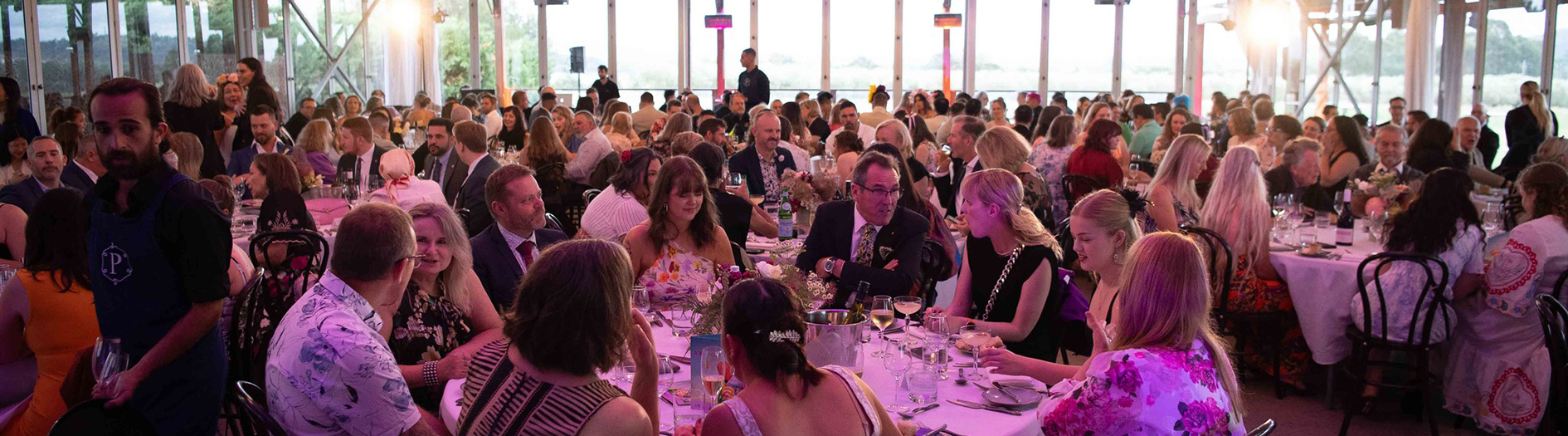 Crowd enjoy meals featuring Canberra cuisine seated at round tables at Pialligo Estate event space, the ceiling is lined with fabric with hanging chandeliers.