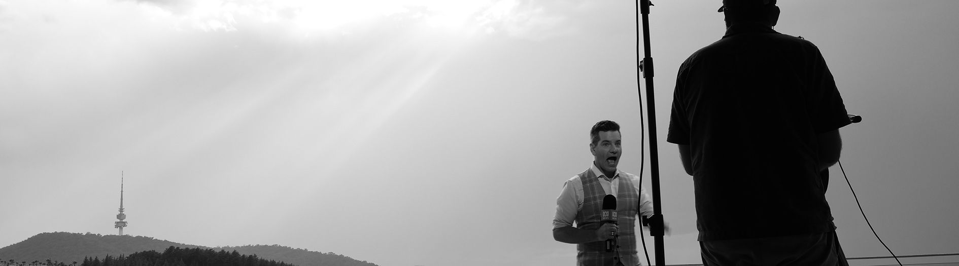 ABC Weather present Nate Byrne stands before a tv camera with the sunlight breaking through a cloudy sky. A dramatic artistic touch has been added by making the image black and white, highlighting the contrast of the sunbeams against the cloudy sky