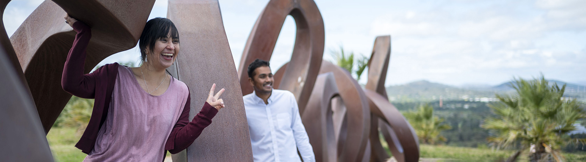 Two adults smile as they lean through the curves of the large metalic 'wide brown land' sculpture